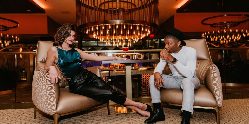 A woman and a man lounging at AZILO Ultra Lounge. Holding cocktails there eyes connect.