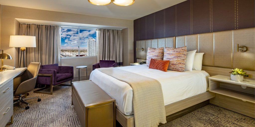 Marra Style King Room. This picture represents the all new Marra Style rooms that are now available to guests. Decor in the room includes a desk, lamp, purple velvet chair, bed with a throw blanket and accessory pillows.
