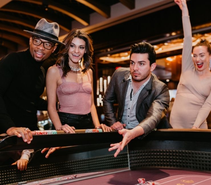 Four people standing around a craps table. One guy is throwing his dice to play while his 3 friends watch. One girl is celebrating with her right arm raised to the roof. Night time casino vibes.