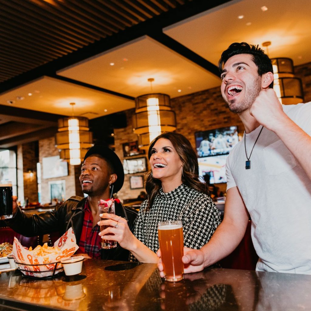 This pictures shows three friends sitting at the bar with drinks and food smiling and cheering because their team just won a sports game on tv